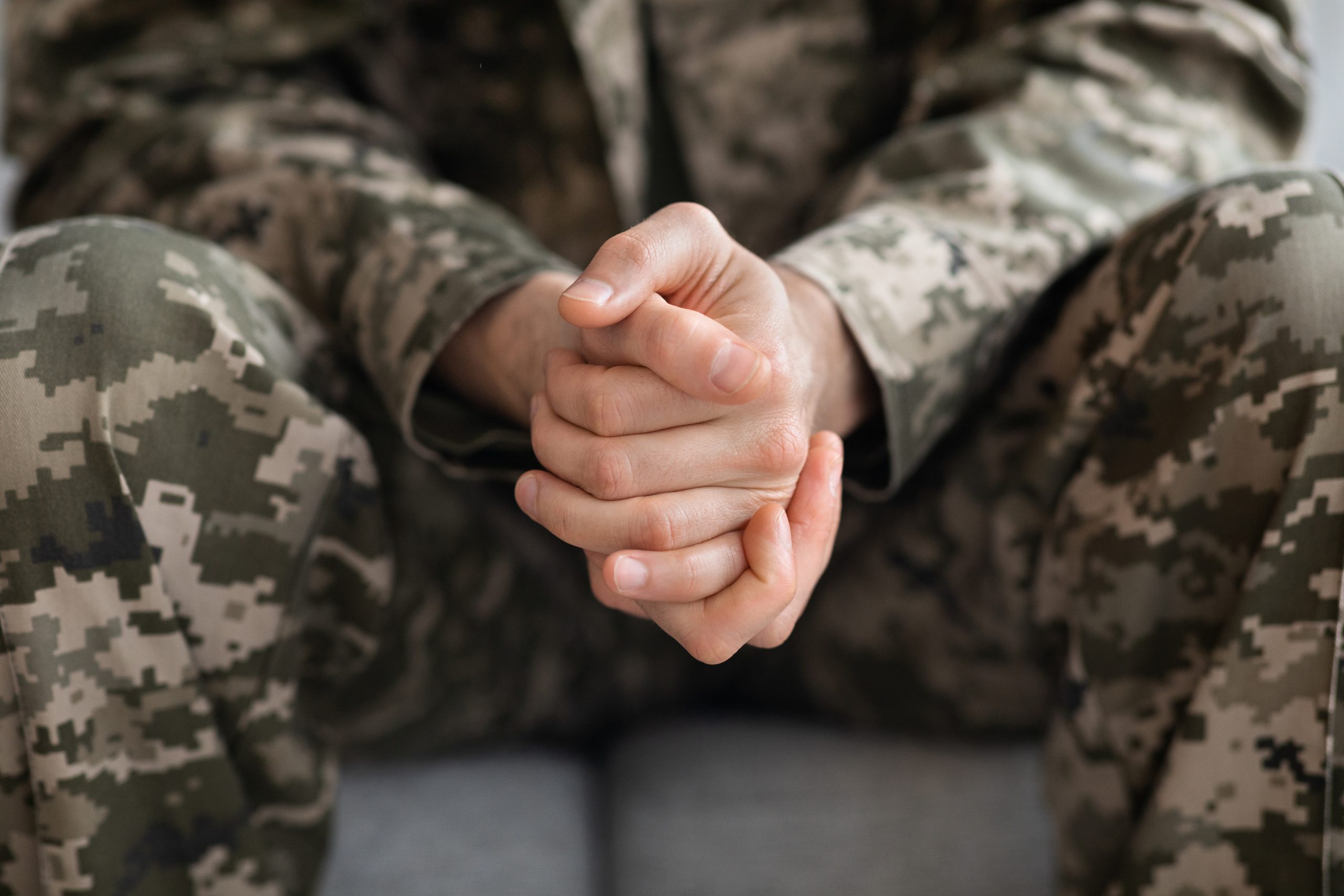 Cropped of soldier sitting on couch alone, suffering from loneliness and depression at home after returning from military service, hands of unrecognizable man in camouflage uniform, closeup